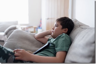 Kid with remote control and looking up with curious face,Young boy sitting on sofa watching cartoon on TV, Portrait Child lying down on couch relaxing in living room after back from school.