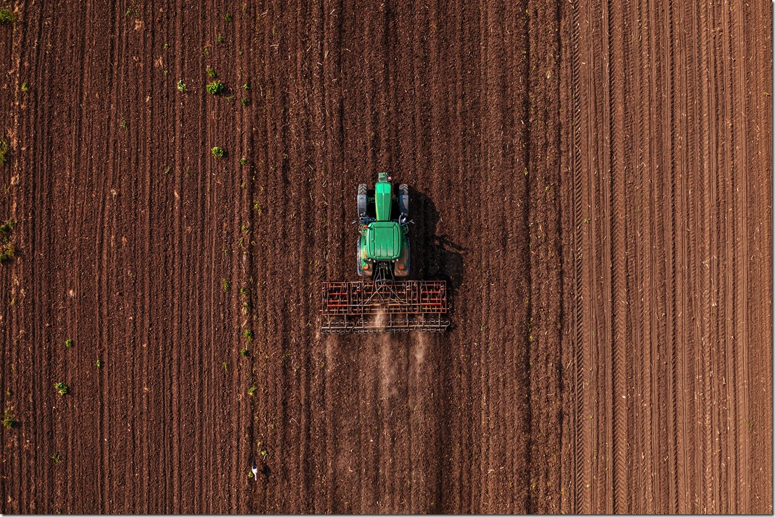 Green tractor vehicle with tiller attached performing field tillage before the sowing season, aerial shot seen from the drone pov