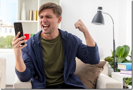 excited young blonde handsome man sits on armchair keeping fist and looking at phone inside design living room