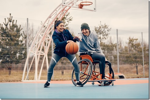 Happy woman and her friend in wheelchair plays basketball on outdoor sports court. 